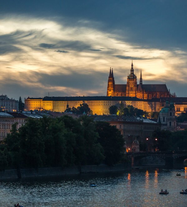 Night view of Prague Castle and the Vltava River with boats at sunset.